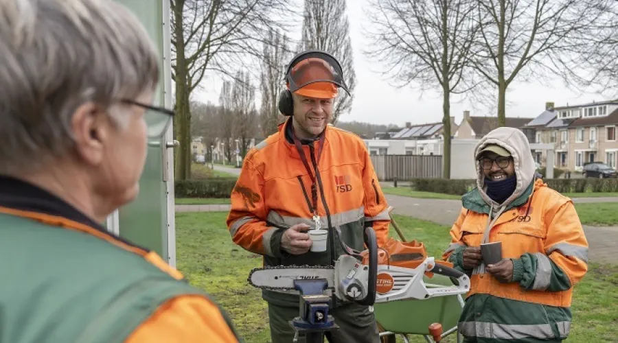 drie groenmedewerkers met elkaar in gesprek met een bakje koffie en een kettingzaag